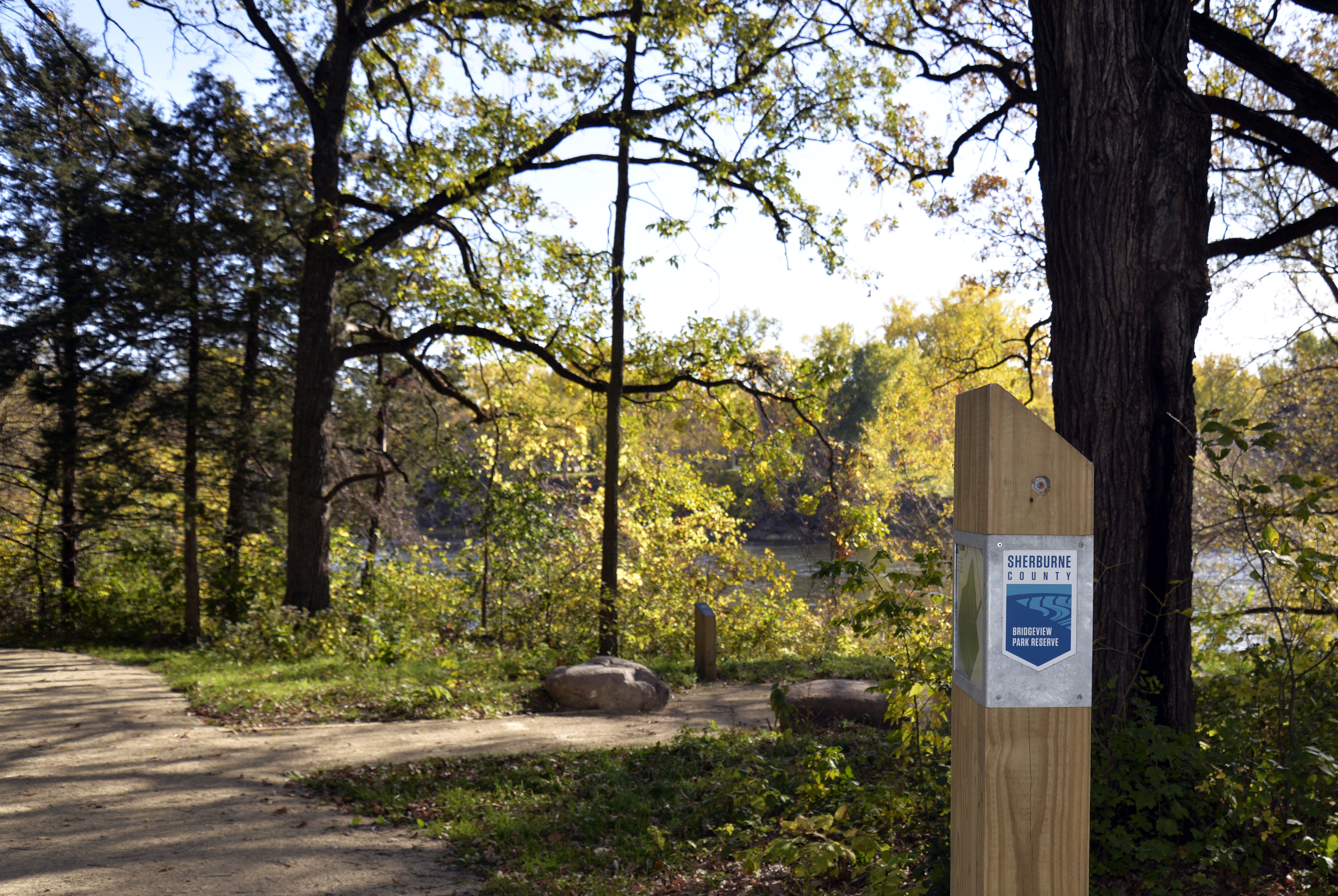 A Sherburne County Parks sign is visible alongside a trail and overlook on the river at Bridgeview Park Reserve.